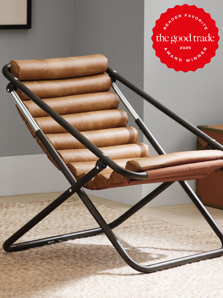 Modern folding chair with a brown cushioned seat and black metal frame in a carpeted room. The top corner has a red badge labeled "The Good Trade Reader Favorite Award Winner 2025.