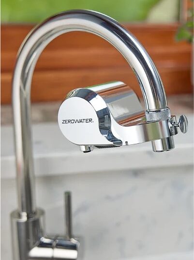 A close-up image of a chrome kitchen faucet with a Zerowater filter attachment installed on the spout. The background shows a marble countertop and a wooden windowsill with greenery.