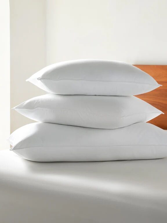 Three white pillows stacked on a bed with a light brown headboard in the background.