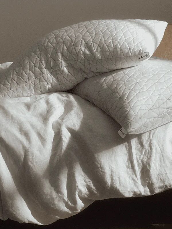 Two white quilted pillows stacked on an unmade bed with white bedding in a softly lit room.
