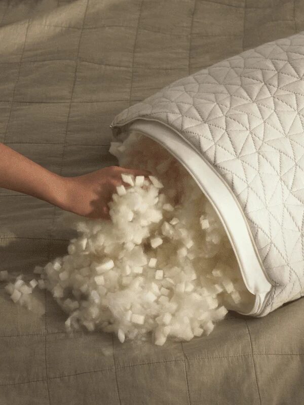 A hand adjusts the stuffing inside a pillow with a quilted cover on a beige bedspread.