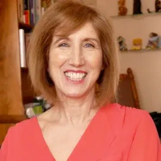 A woman with shoulder-length brown hair smiles while wearing a coral top. Shelves and a wooden bookcase are in the background.