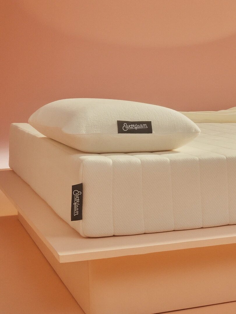A white mattress with a matching pillow on a minimalist, light pink bed frame against a soft pink background. Tags on both the mattress and pillow read "Eucalyptus.