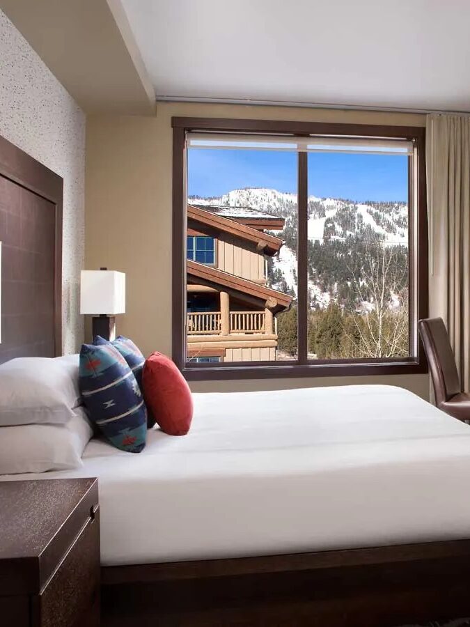 A hotel room with a large bed, desk, chair, and TV. The window offers a scenic view of snowy mountains and a building outside. The room has beige walls and dark wood furnishings.
