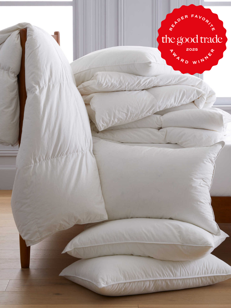 A stack of white pillows and comforters piled on a chair. Wooden floor and window in the background. A red "The Good Trade 2025 Reader Favorite Award Winner" badge is displayed.