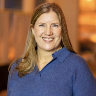 A woman with long, light brown hair is smiling at the camera. She is wearing a blue sweater and standing indoors with a blurred background.