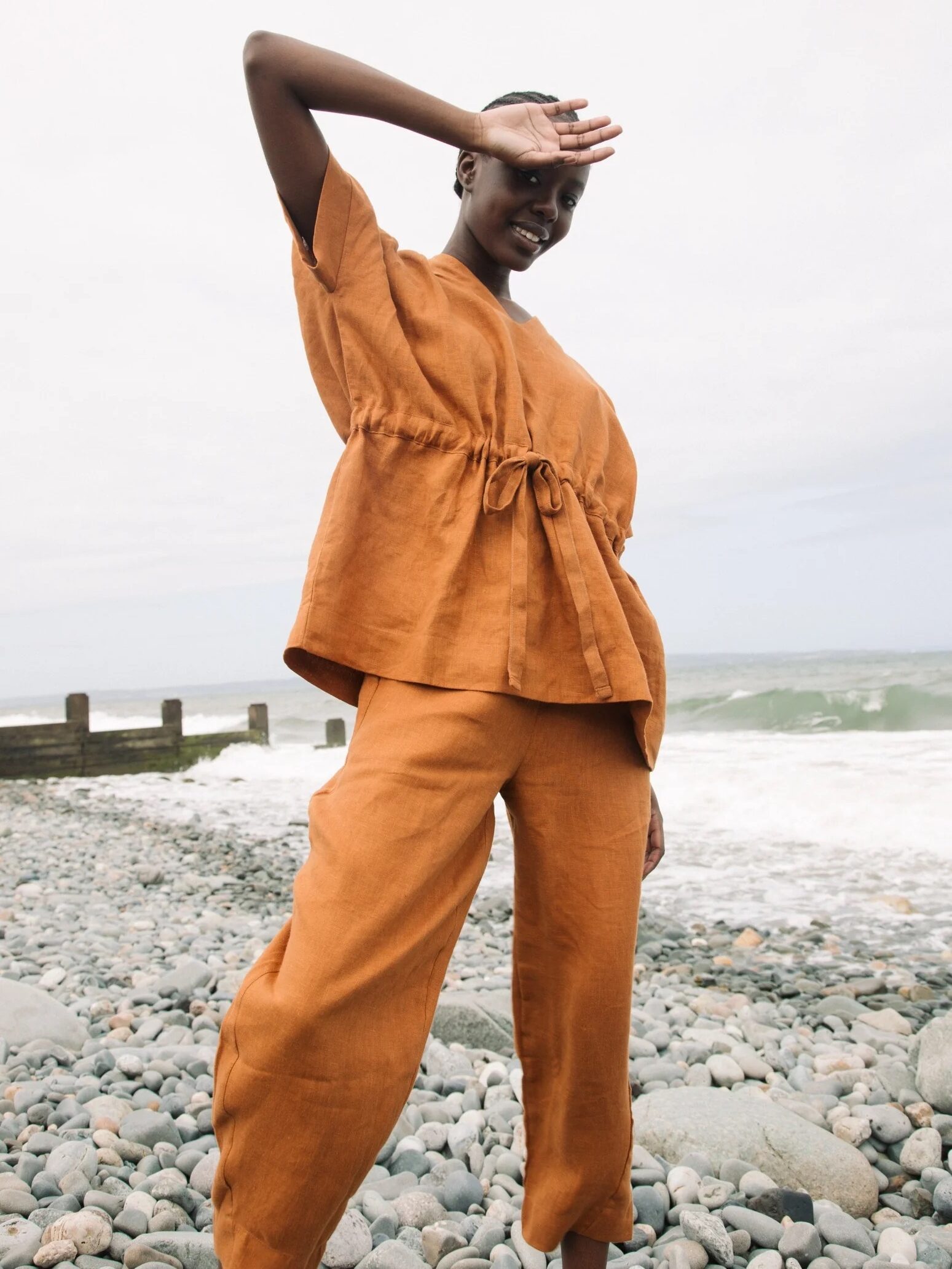 A person in orange clothing stands on a rocky beach, shielding their eyes from the sun with one hand. The ocean and a cloudy sky are visible in the background.