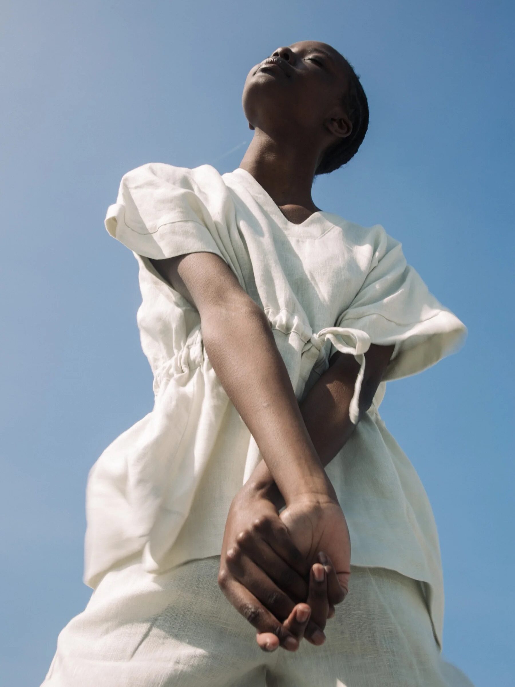 A person wearing a white outfit stands against a clear blue sky with their hands clasped and arms extended downwards, gazing upwards.