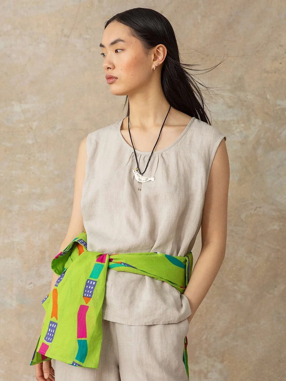 A person wearing a sleeveless beige top, matching beige pants, a green scarf with colorful patterns tied around the waist, and a necklace stands with their head turned to the side.