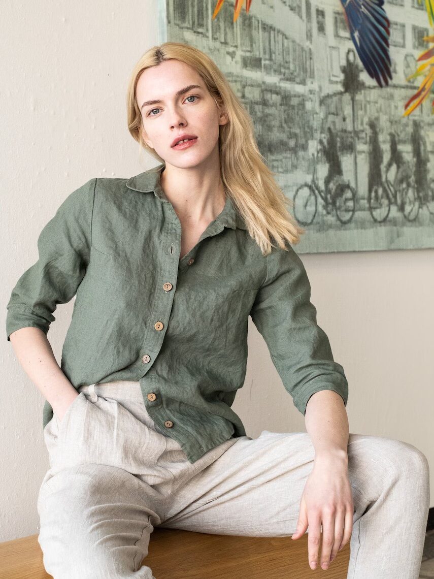 A person with blonde hair is sitting on a wooden surface, leaning back slightly, wearing a green button-up shirt and light-colored pants. A colorful wall mural with bicycles is visible in the background.