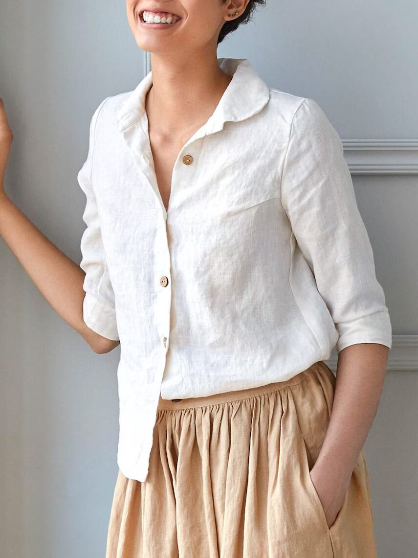 Person smiling, wearing a white button-up shirt and a beige skirt, standing with one hand resting on a wall.