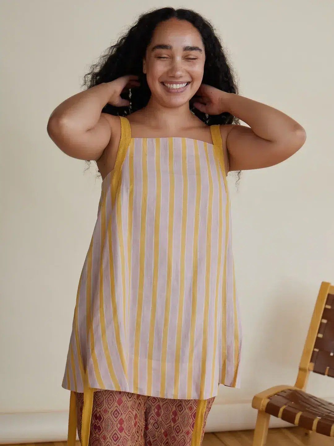 A person with long curly hair smiles with eyes closed, wearing a sleeveless striped top and patterned pants, standing near a woven chair.