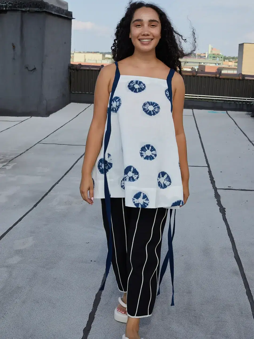 A person with curly hair smiles and walks on a rooftop. They are wearing a sleeveless top with circular patterns and striped pants.