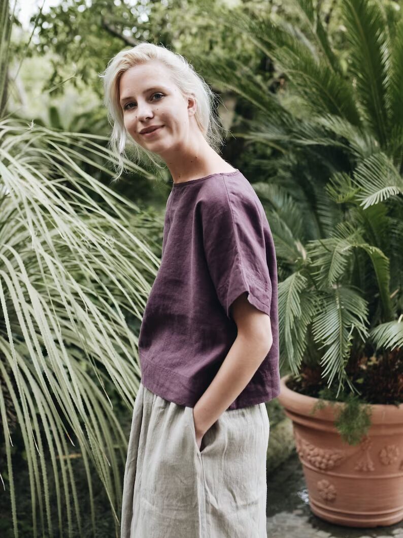 A person with light hair stands outdoors, wearing a purple top and beige pants. They are in a garden with various green plants and trees in the background.
