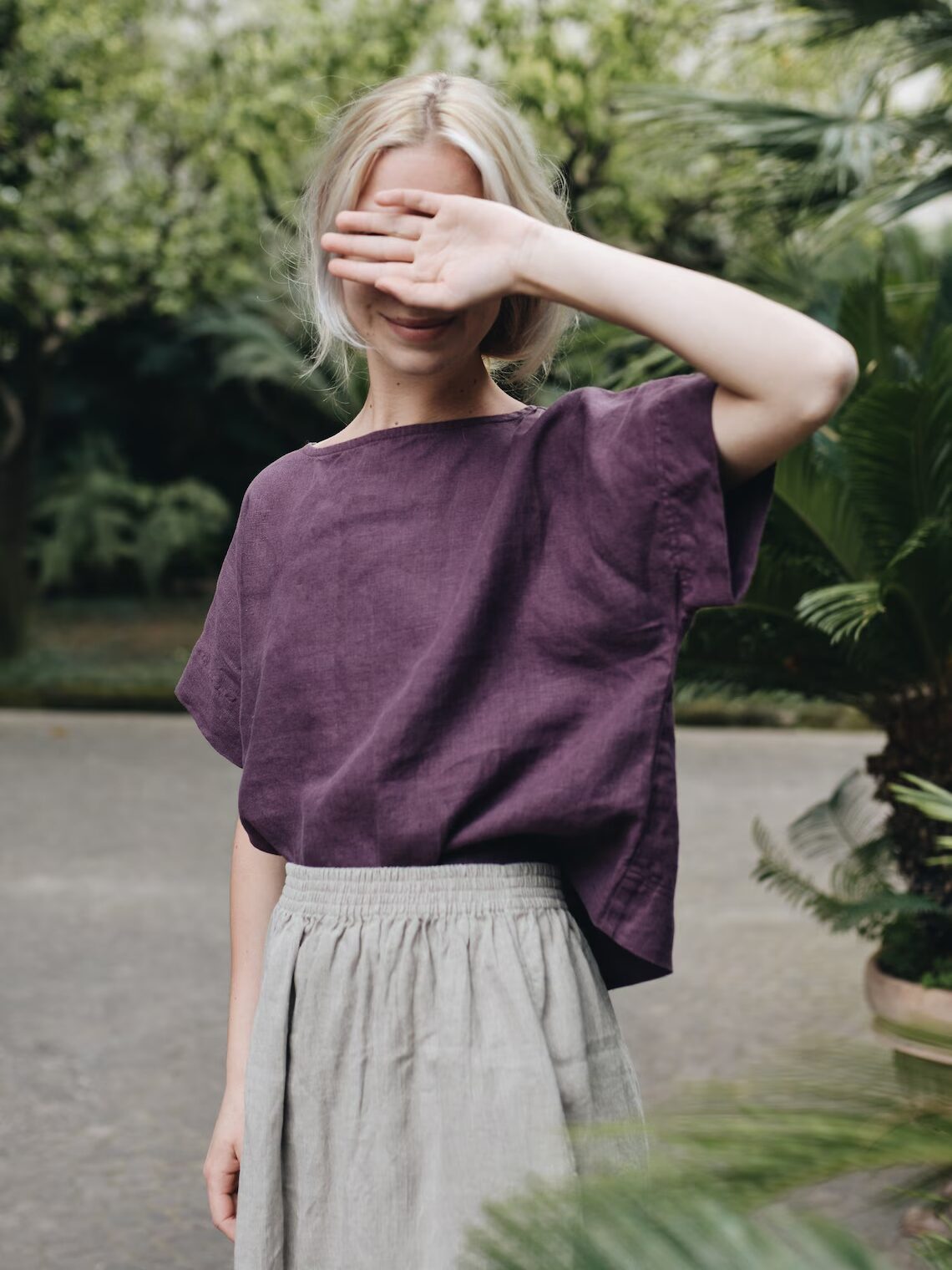 A person with blonde hair stands outside, covering their face with one hand. They wear a purple shirt and light gray skirt, surrounded by green plants.