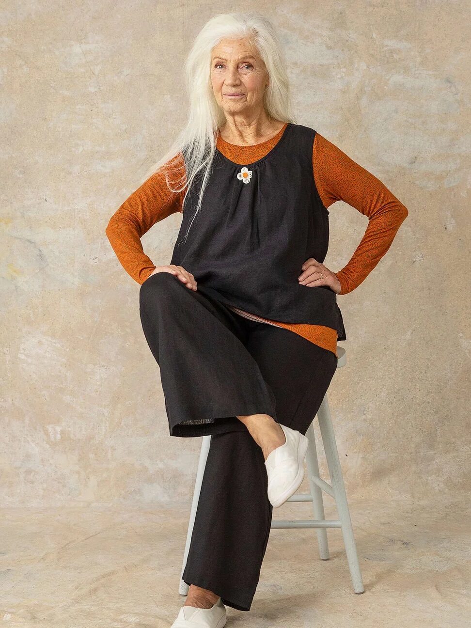 Elderly woman with long white hair sits on a stool, wearing an orange long-sleeve shirt, black sleeveless tunic, black pants, and white shoes. The background is a textured beige wall.