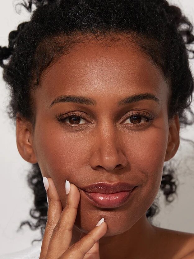 A person with curly hair and natural makeup touching their face with their right hand, displaying clear skin and white manicure. The background is neutral.