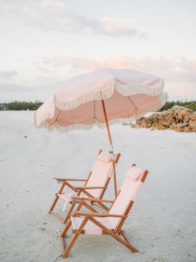 Two wooden lounge chairs with pink and white striped fabric sit under a matching fringed beach umbrella on a sandy beach. Rocks and a few clouds are visible in the background.