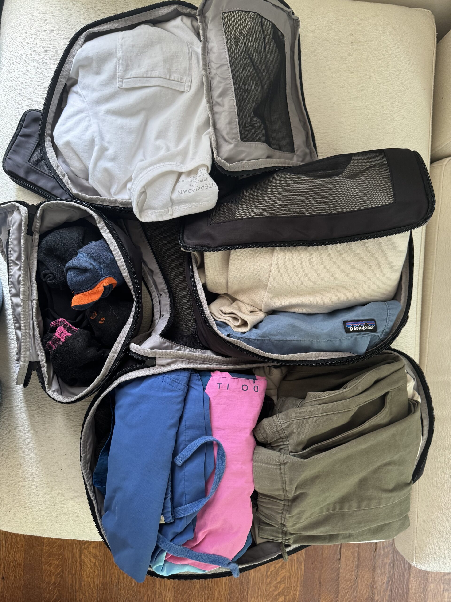 Open, organized packing cubes filled with neatly folded clothing items, including shirts, pants, and socks, laid out on a light-colored surface.