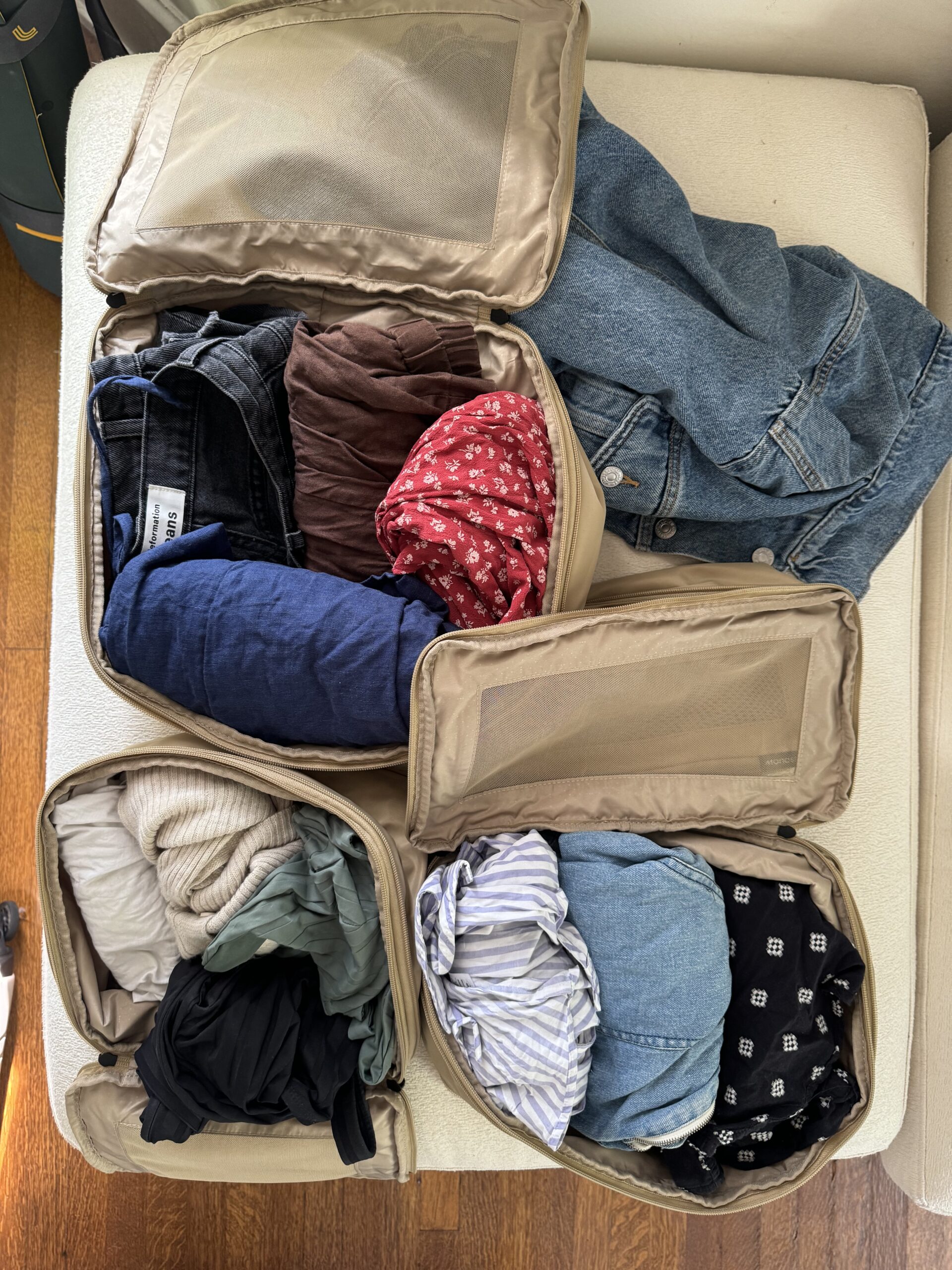 An organized arrangement of clothes in beige packing cubes and a denim jacket placed on a white surface.