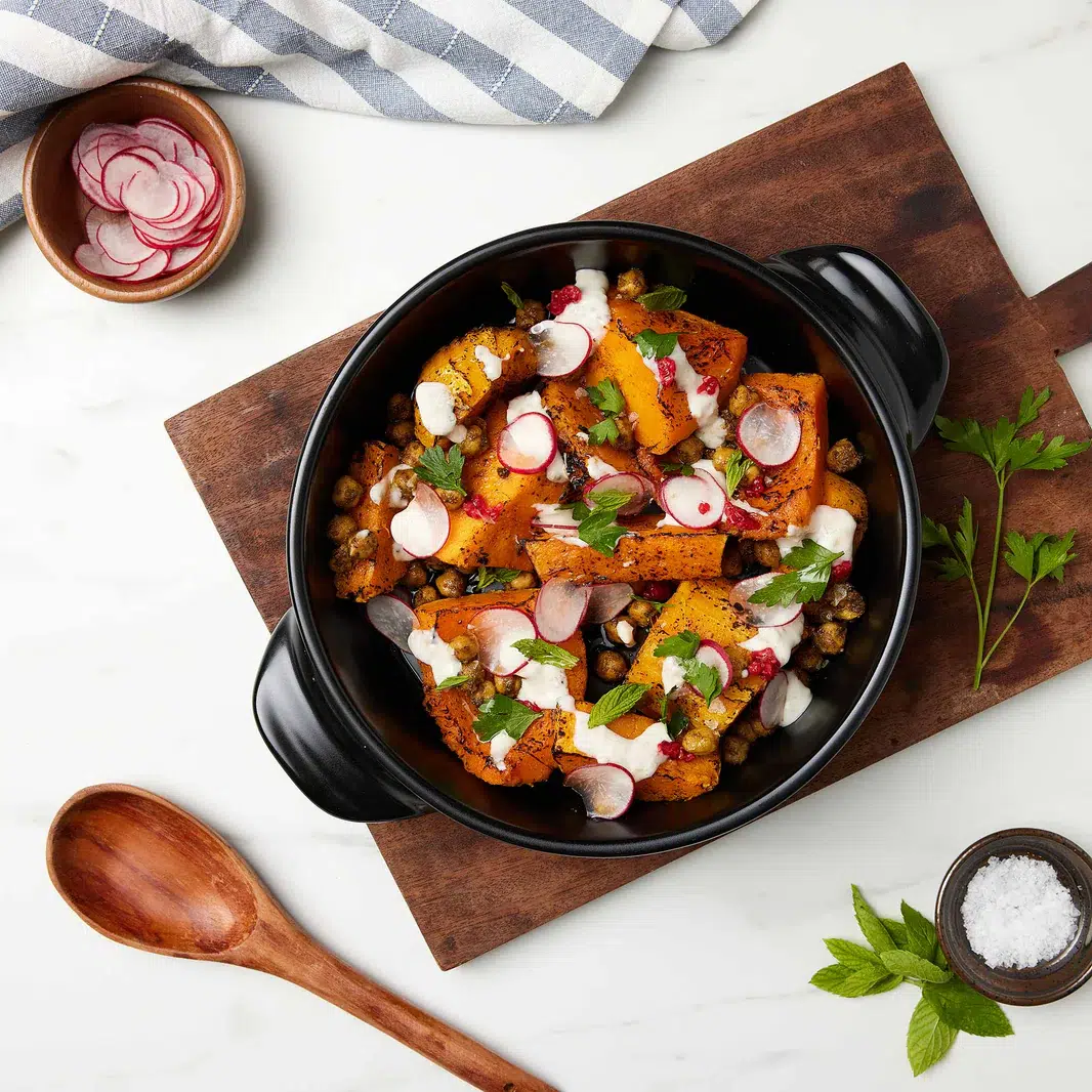 A black bowl filled with roasted vegetables, chickpeas, and radish slices on a wooden board. Garnished with herbs and yogurt. Accompanied by a wooden spoon, a small bowl of radishes, herbs, and salt.