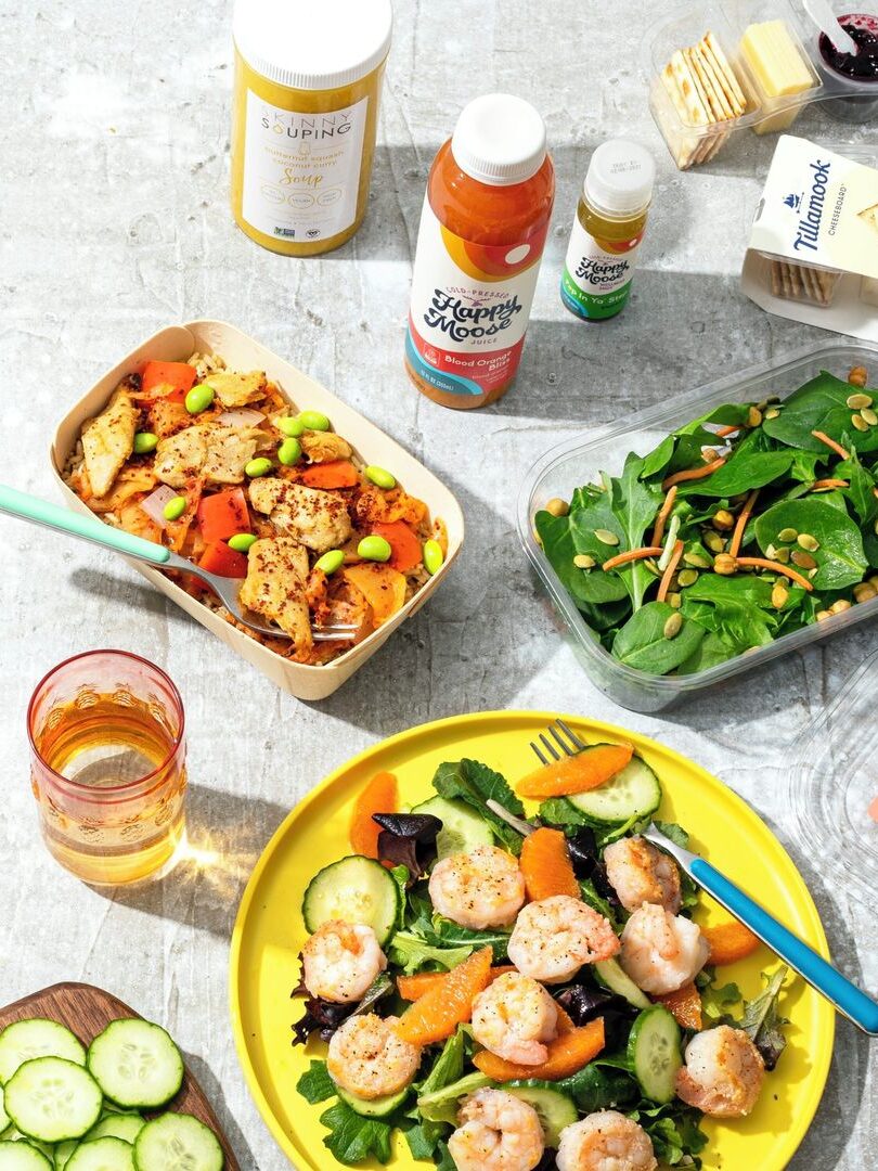 A variety of food items are arranged on a surface, including a salad with shrimp, a vegetable dish, spinach salad, cheese, and beverage bottles. A fork and a glass of drink are also visible.