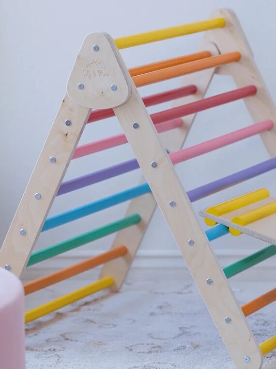 A toddler wearing a headband sits on a wooden climbing structure with colorful rungs and holds onto a sloped ramp. The structure is branded as "Little Climber.