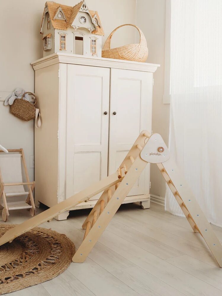 A child’s room with a wooden play set, white cabinet, wicker basket, and dollhouse on top. Sunlight filters through a sheer curtain, illuminating light wood floors.