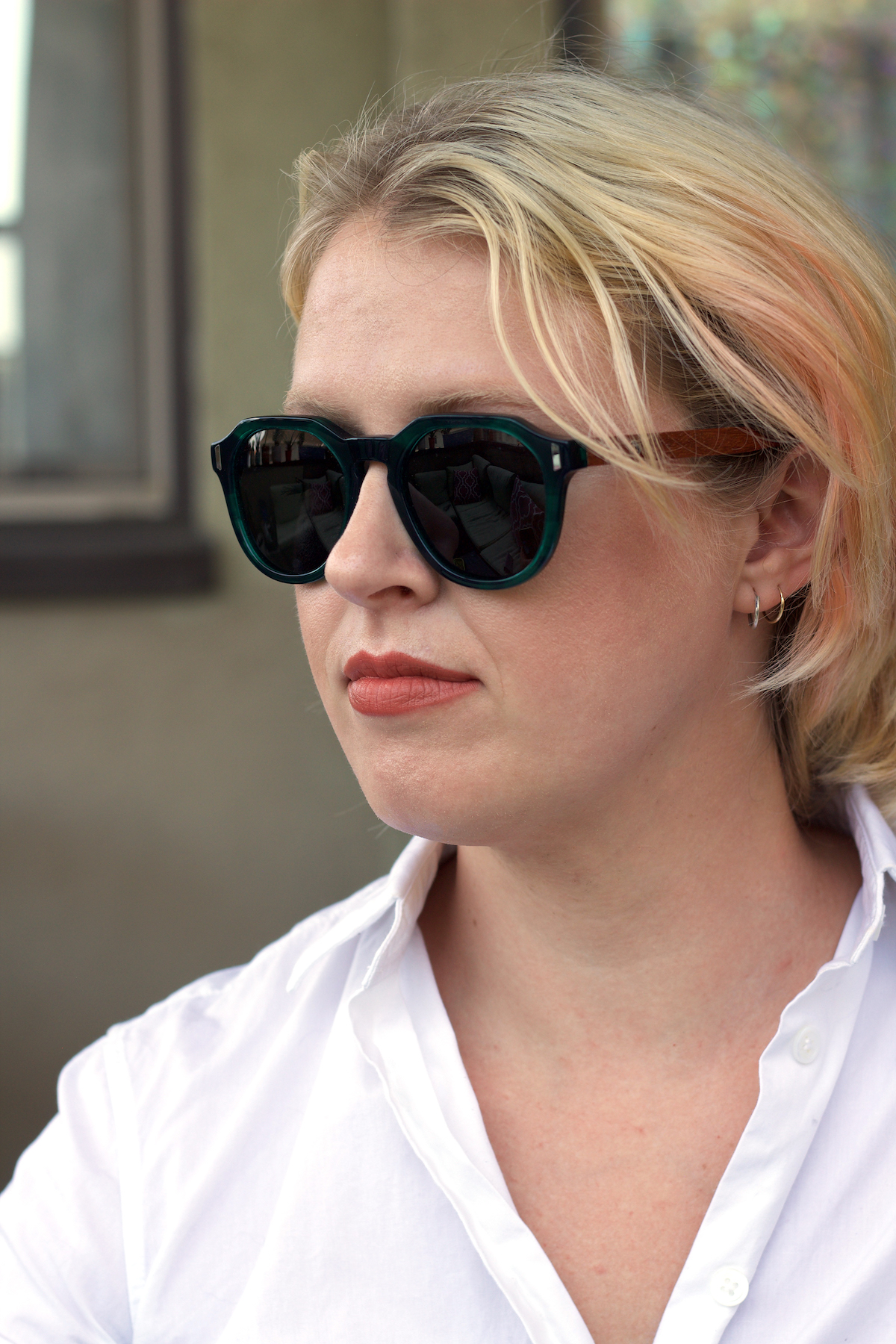 A woman wearing dark sunglasses and a white collared shirt is seen in a close-up, outdoors setting.