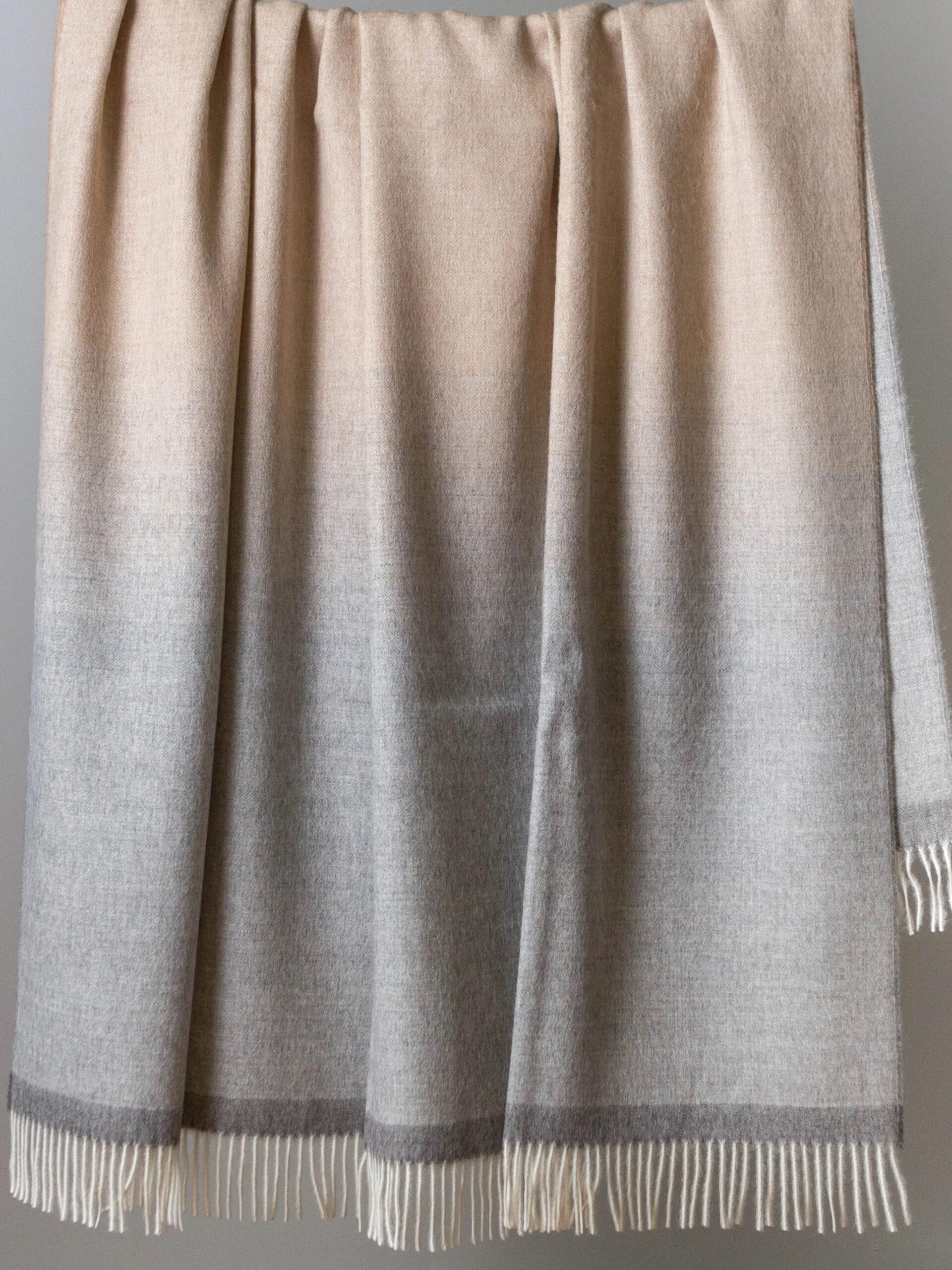 A beige and light gray woolen shawl with a gradient design is draped on a surface. The shawl ends in fringes.