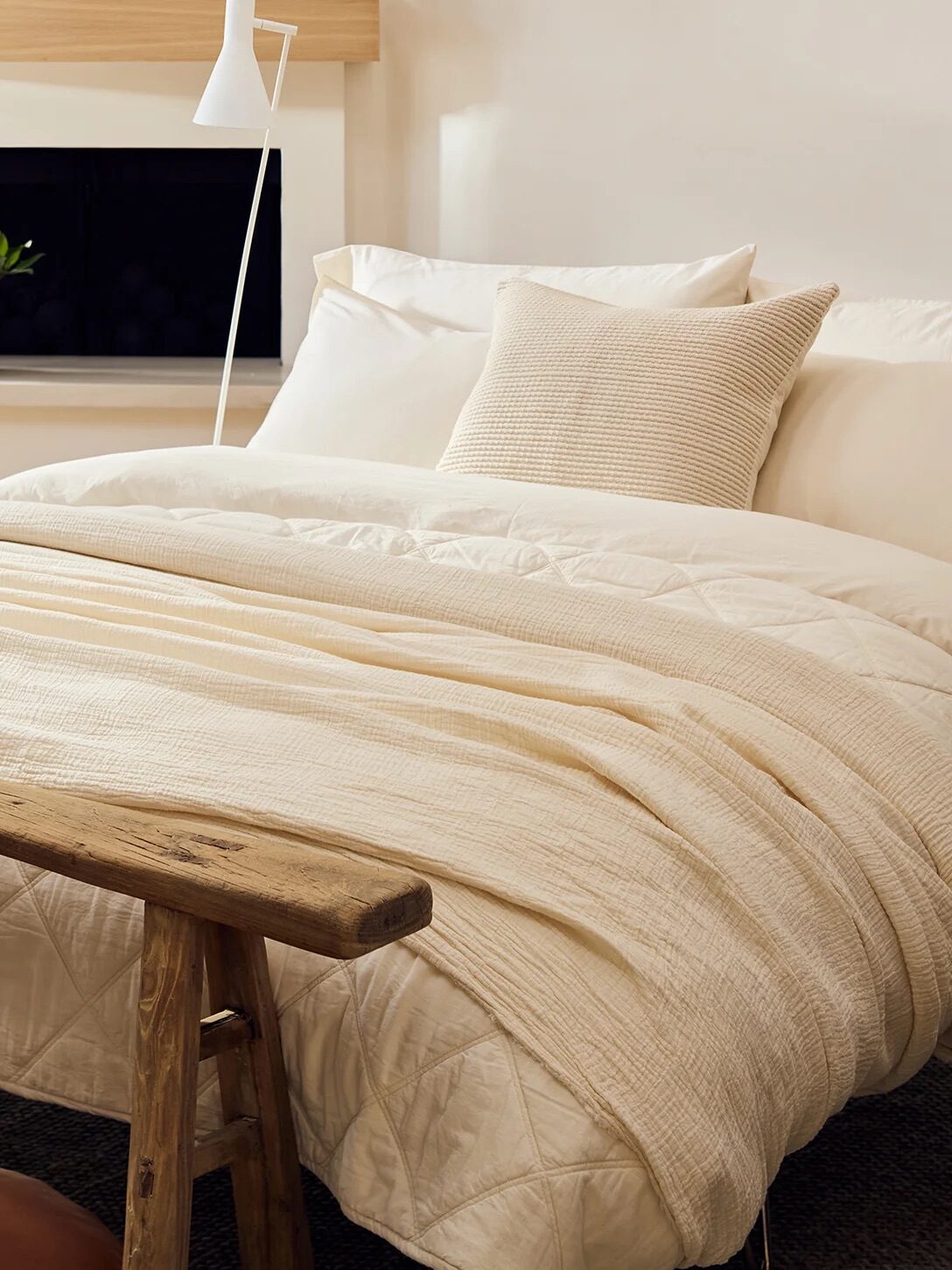 A neatly made bed with cream-colored bedding is in a bright room featuring a wooden bench at the foot of the bed, a potted plant on the bench, and a flowering plant on a shelf above the bed.