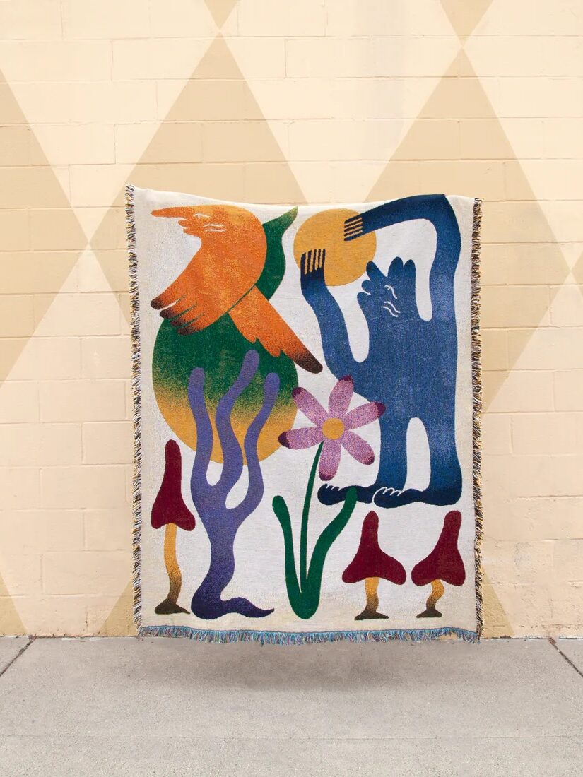A woven blanket featuring abstract, colorful illustrations of a figure, a bird, a flower, mushrooms, and plant-like shapes, displayed against a beige wall with a geometric pattern.