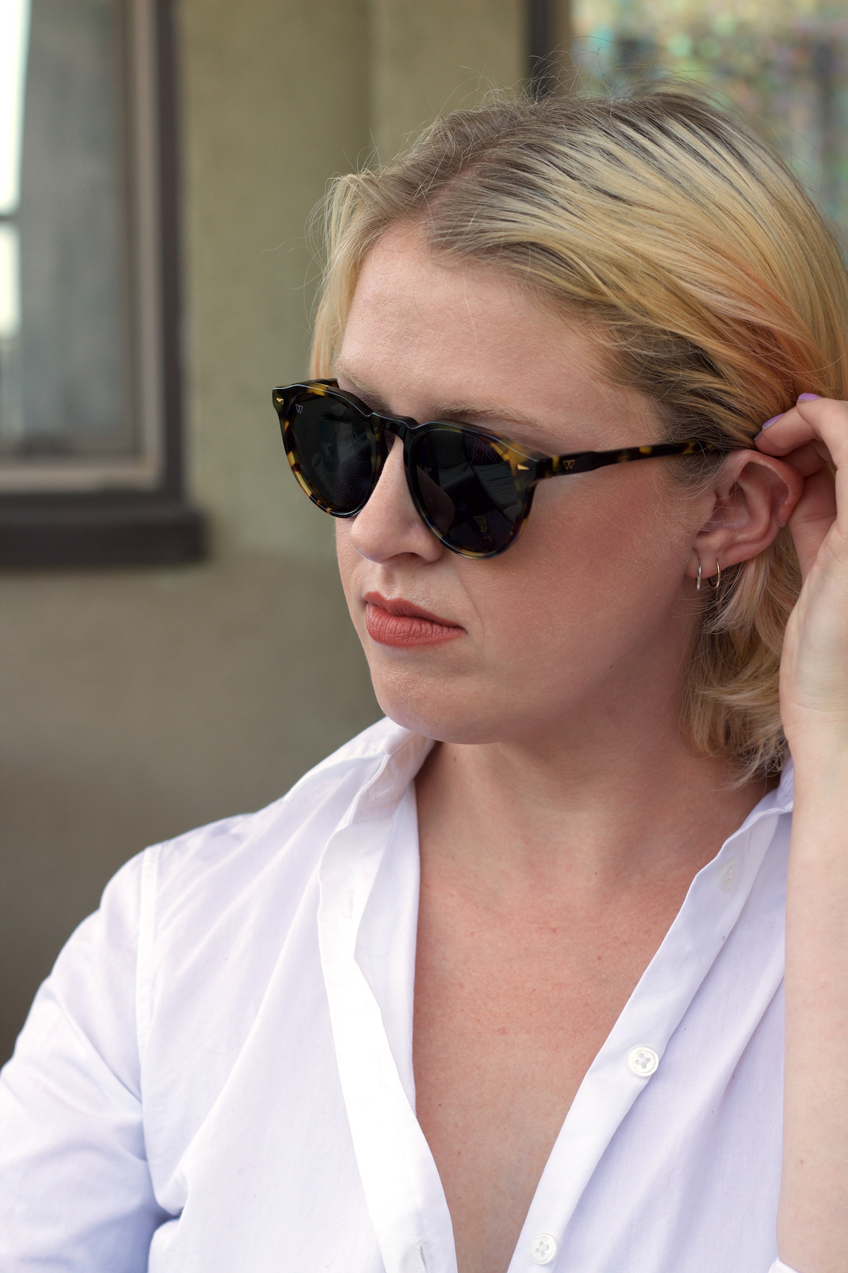 A person with blonde hair wearing black sunglasses and a white shirt is looking to the side while touching their hair.