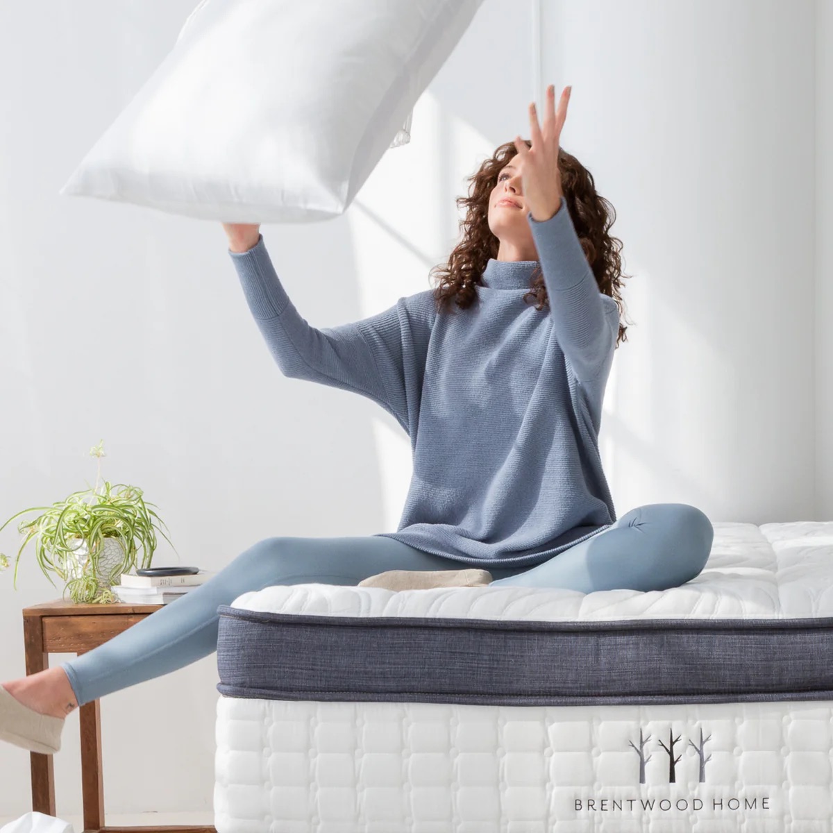 A woman sitting on a bed tosses a pillow in the air. The room is bright with natural light, and there is a small plant on a side table next to the bed.