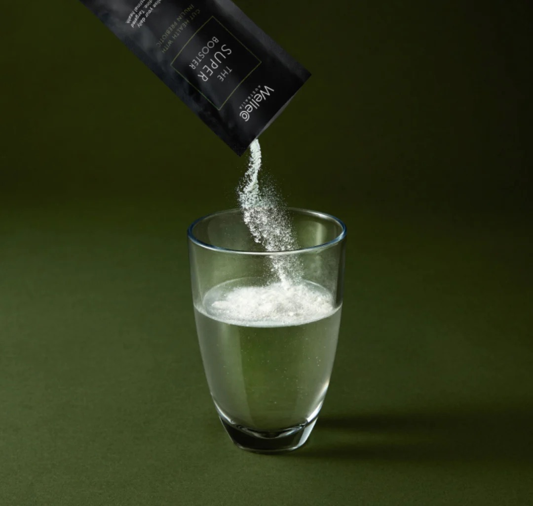 A packet labeled "Super Booster" is being poured into a glass of water against a dark green background.