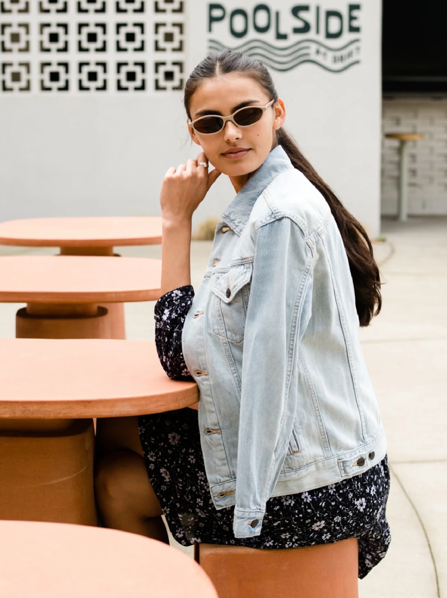 A woman wearing sunglasses, a light denim jacket, and a black floral dress sits at an outdoor table in front of a wall with a "Poolside" sign.
