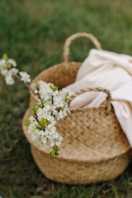 A woven basket with a light cloth and a branch of white blossoms rests on green grass.