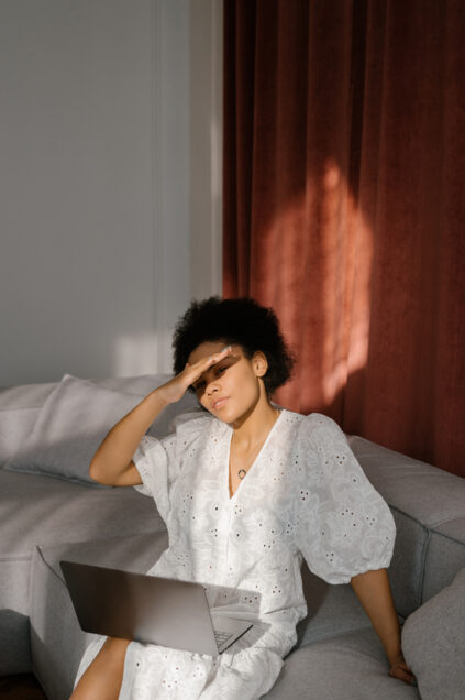 A person in a white dress sits on a couch with a laptop, shielding their eyes with one hand.