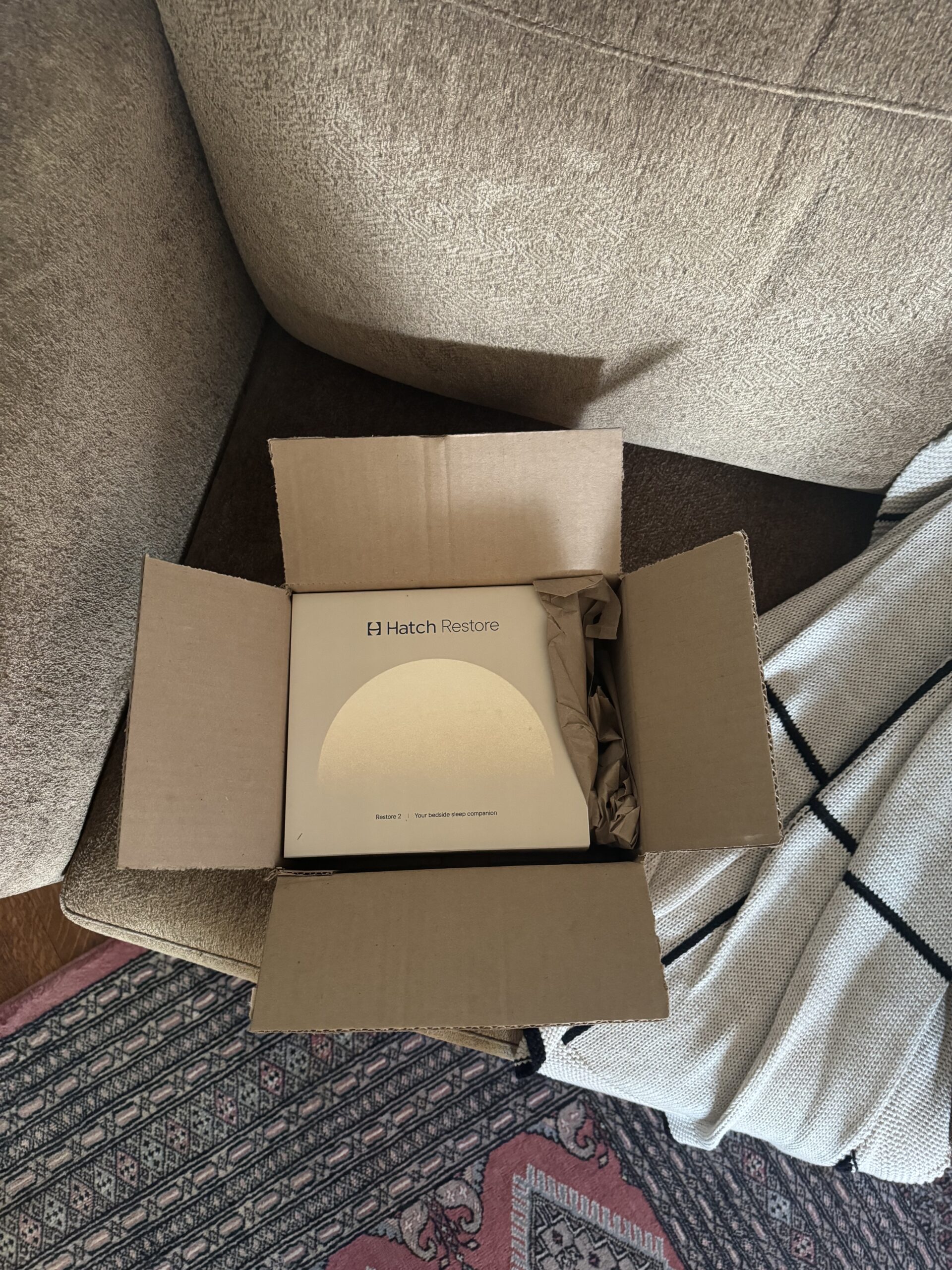 Open cardboard box containing a Hatch Restore device, placed on a beige sofa next to a black-and-white blanket.