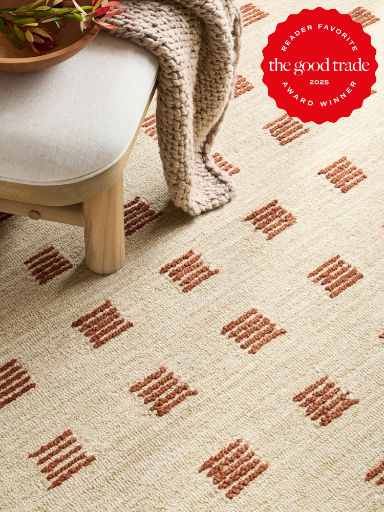 Beige woven rug with a red geometric pattern, beside a light wood chair with a cream cushion and a chunky knit beige throw. "The Good Trade 2025 Award Winner" badge appears in the corner.