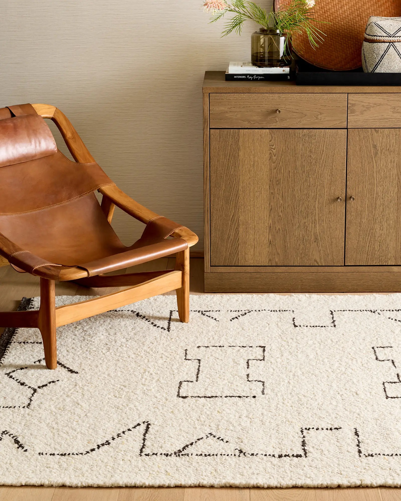 A wooden armchair with a leather seat sits next to a wooden cabinet on a white rug with a geometric black pattern. A vase with flowers and woven baskets are on the cabinet.