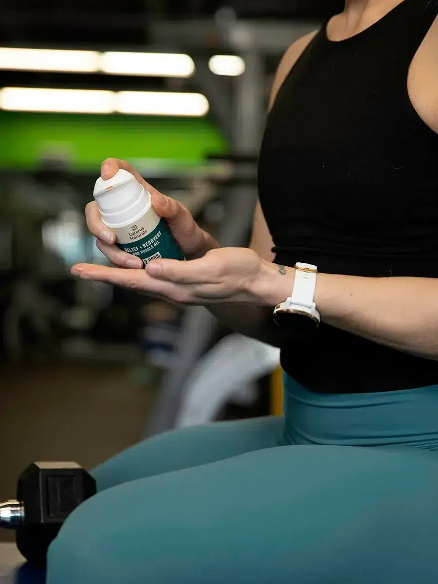 Person in workout attire applying a cream from a bottle onto their hand, seated in a gym setting with exercise equipment visible in the background.