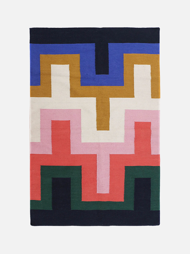 Rectangular rug with a geometric, zigzag pattern in blue, mustard yellow, pink, red, green, black, and white on a white background.