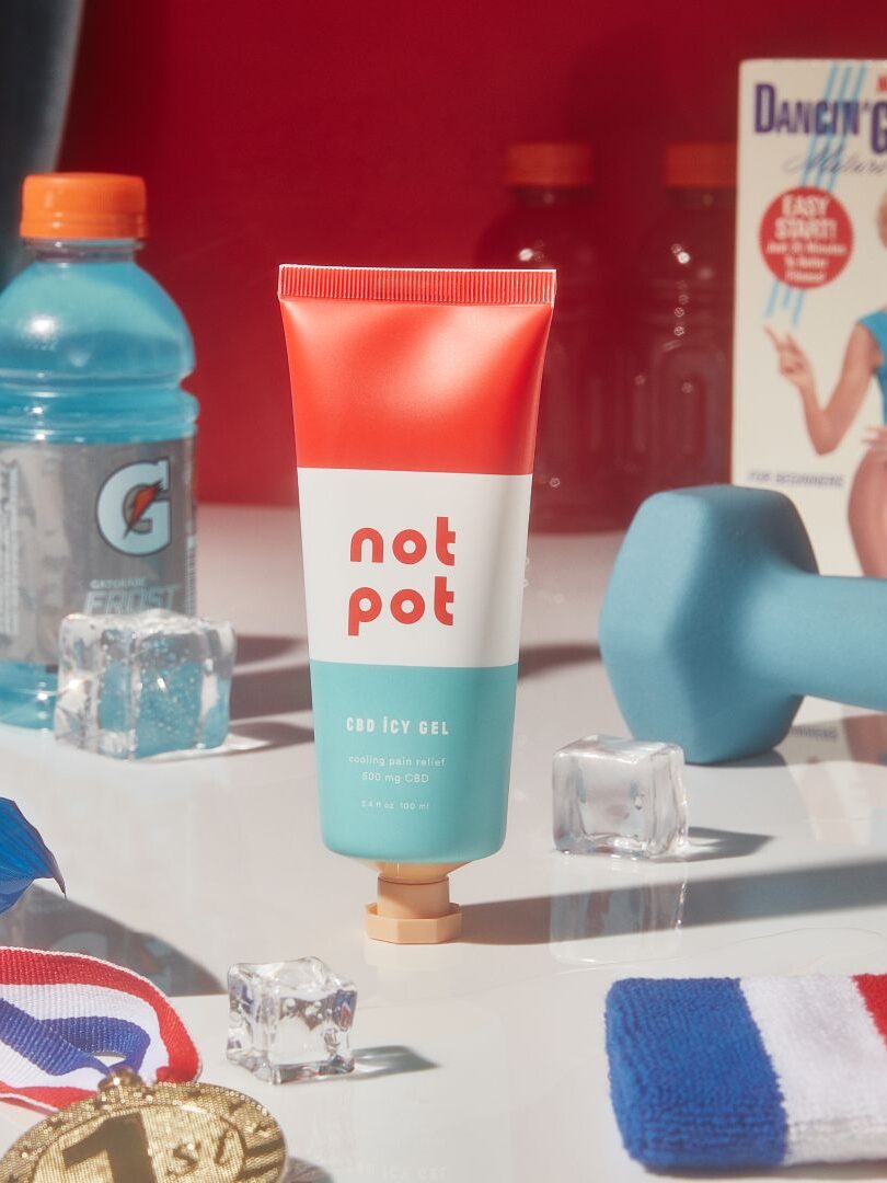 A tube of Not Pot CBD Icy Gel is placed on a surface surrounded by Gatorade, a dumbbell, ice cubes, a medal, an 80s exercise VHS tape, and a sports bottle.