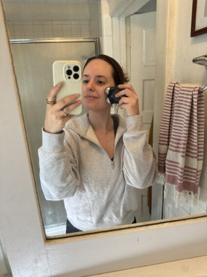 A person stands in a bathroom, holding a phone with a white case to take a selfie while using a shaving device on their neck with their other hand. They wear a light grey sweatshirt.