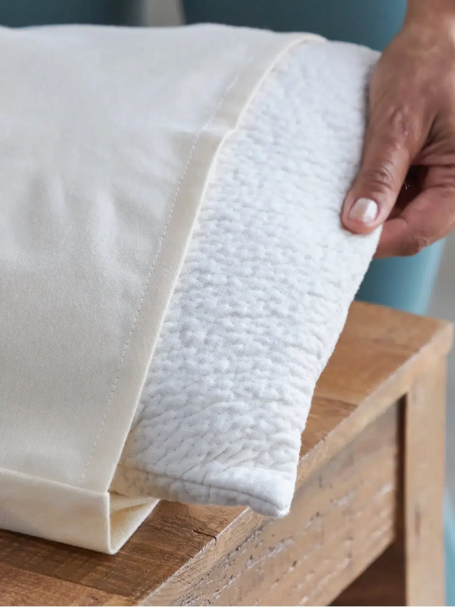 A hand holding the corner of a white blanket with a soft, textured interior, partially folded over a wooden table.