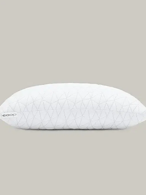 A white, quilted pillow with a tag labeled "Coop" against a plain, beige background.