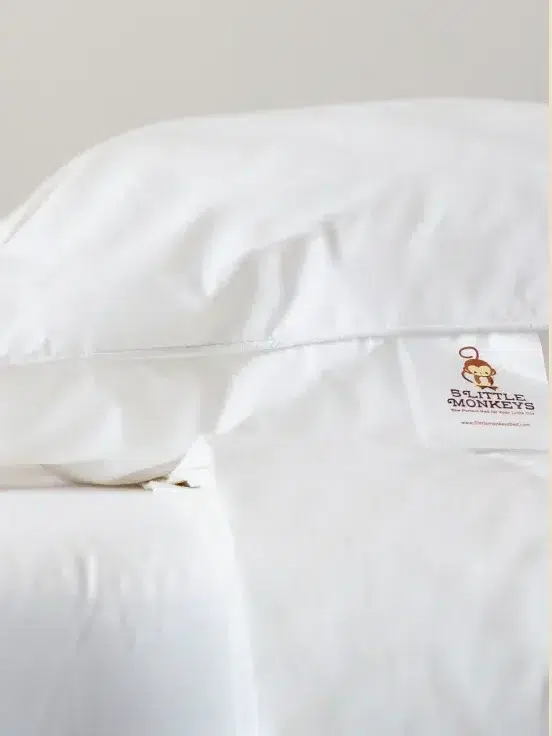 A stuffed monkey toy sits on a neatly made bed beside white pillows and bedding.