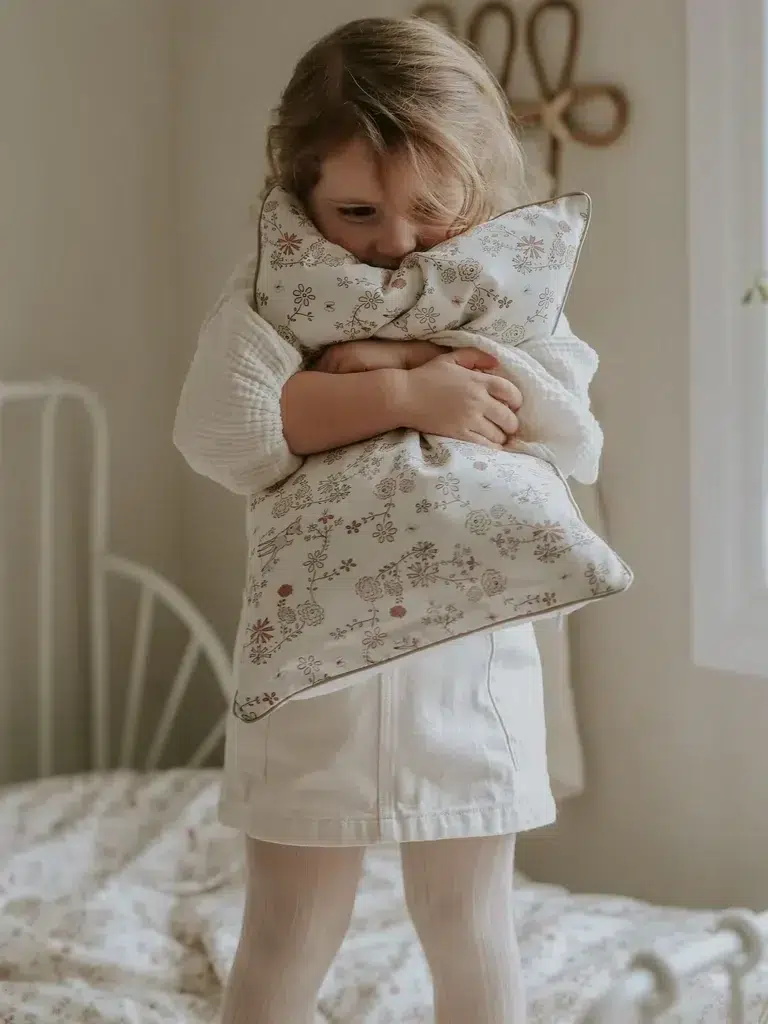 A young child stands on a bed, holding a patterned pillow close to their face. The room has soft lighting, a white bed frame, and a window with a flower vase on the sill.
