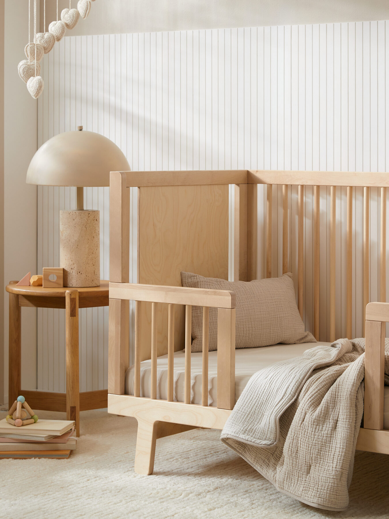 A cozy nursery with a wooden crib, a side table with a lamp, and a small rattan chair with stuffed toys. The room has light walls, a soft rug, and large window allowing natural light in.